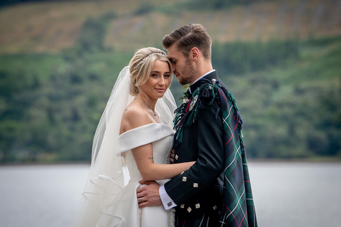 A groom wearing a tartan fly plaid over a black jacket with silver buttons nuzzles into a bride wearing an off-shoulder wedding dress. The landscape and water in the background are soft focus