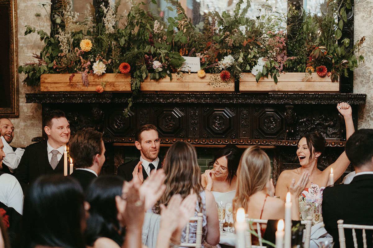 Bride and groom react during wedding speeches at the top table, framed by abundant seasonal floral arrangements and candlelight at Cambo Country House.