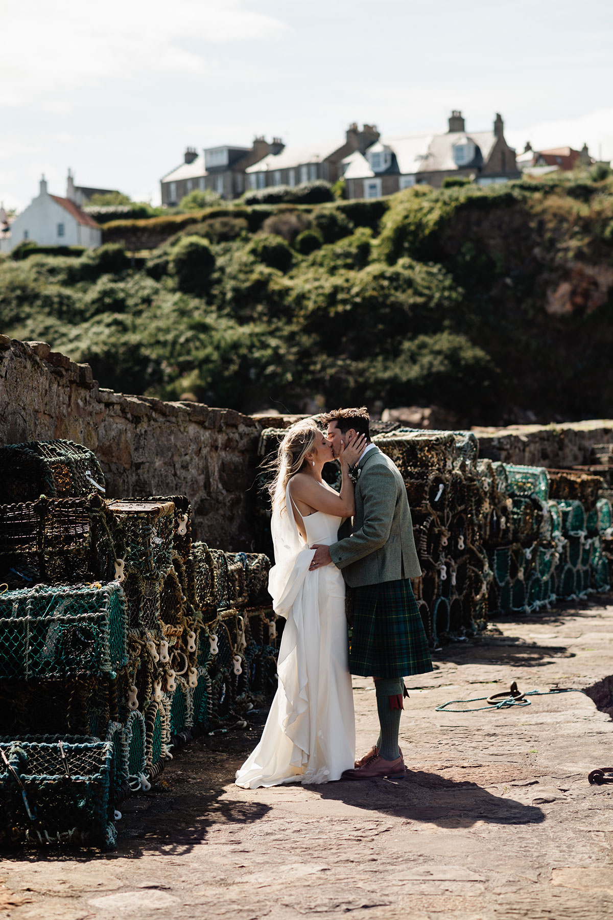Bride and groom kissing beside stacks of fishing creels at the harbour on a sunny day.