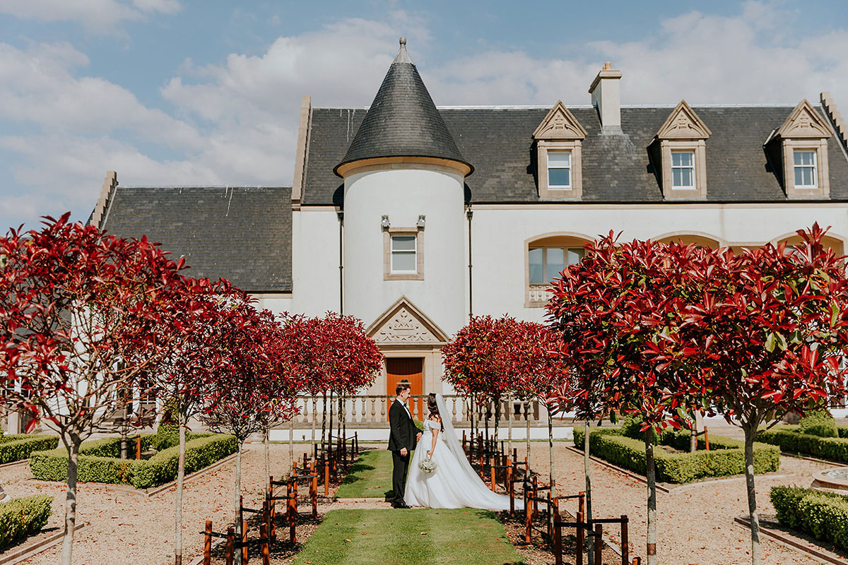 bride and groom stand on grass walkway underneath red trees at ingliston country club