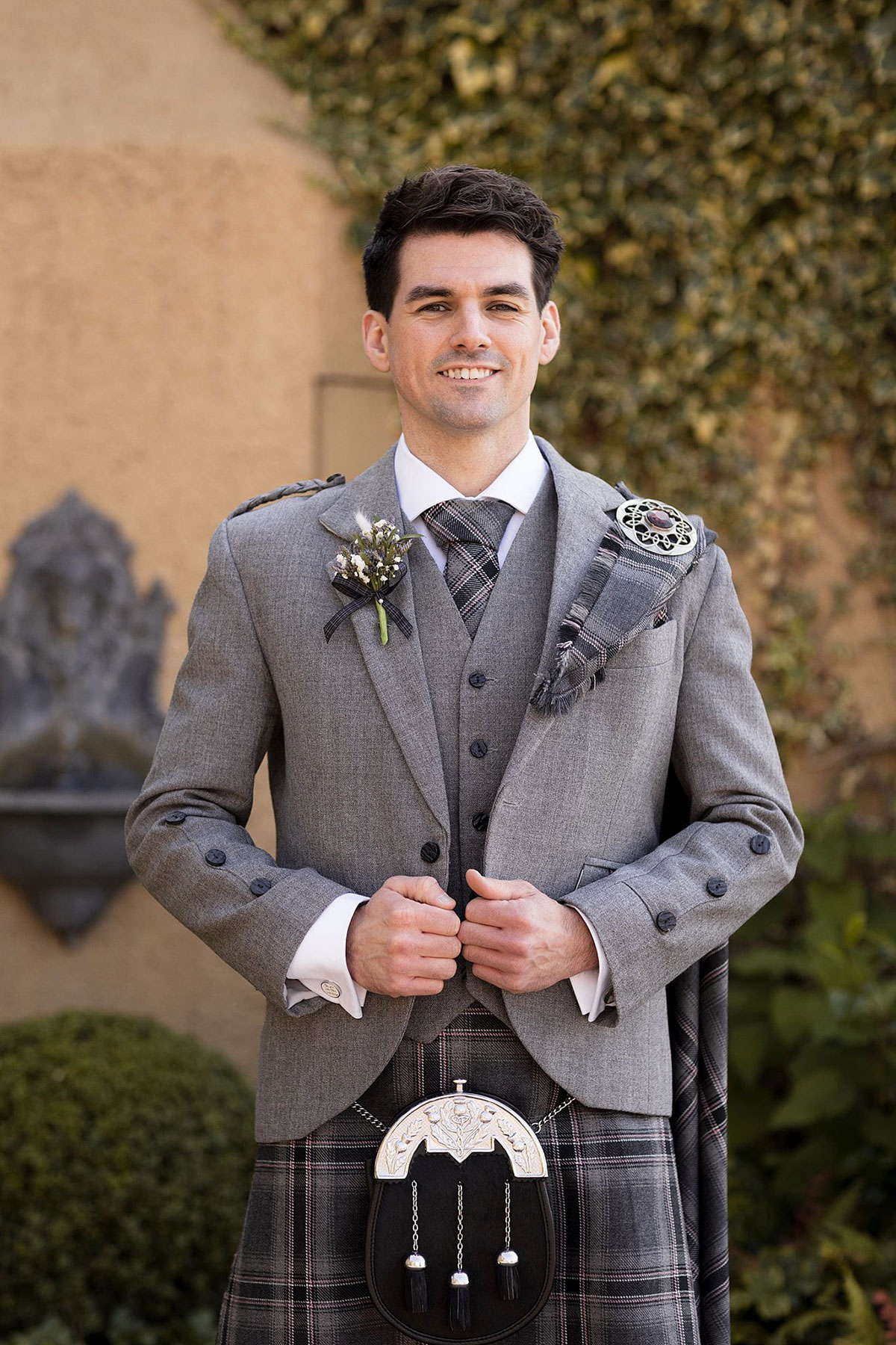 Groom wearing grey Scottish kilt outfit and sporran posing outside Old Course Hotel during St Andrews wedding.