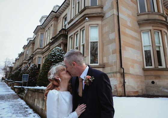 a man and woman kiss in front of yellow sandstone building with snow on the ground 