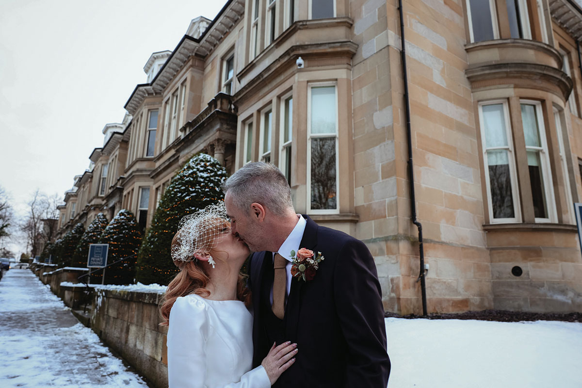 a man and woman kiss in front of yellow sandstone building with snow on the ground