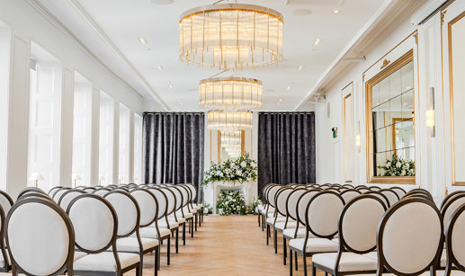 A long room set up for a wedding ceremony with white round-back chairs on either side, chandeliers along the centre of the roof and white flowers on at the end of the aisle