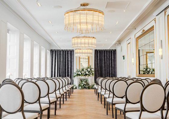 A long room set up for a wedding ceremony with white round-back chairs on either side, chandeliers along the centre of the roof and white flowers on at the end of the aisle