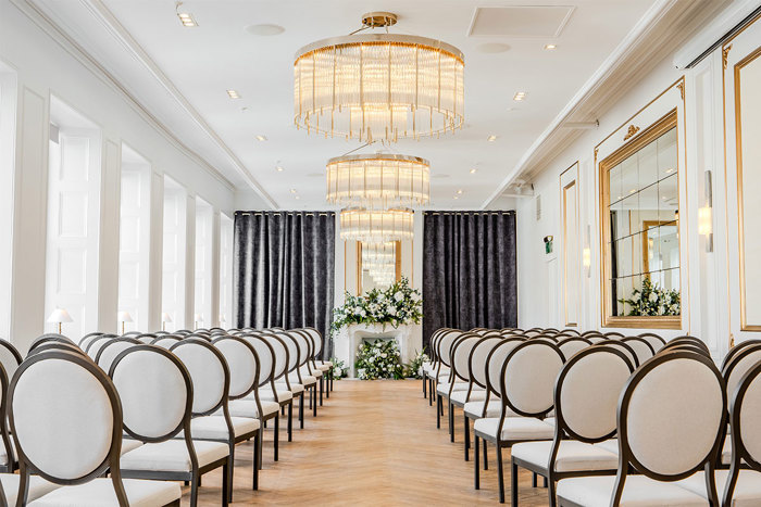 A long room set up for a wedding ceremony with white round-back chairs on either side, chandeliers along the centre of the roof and white flowers on at the end of the aisle