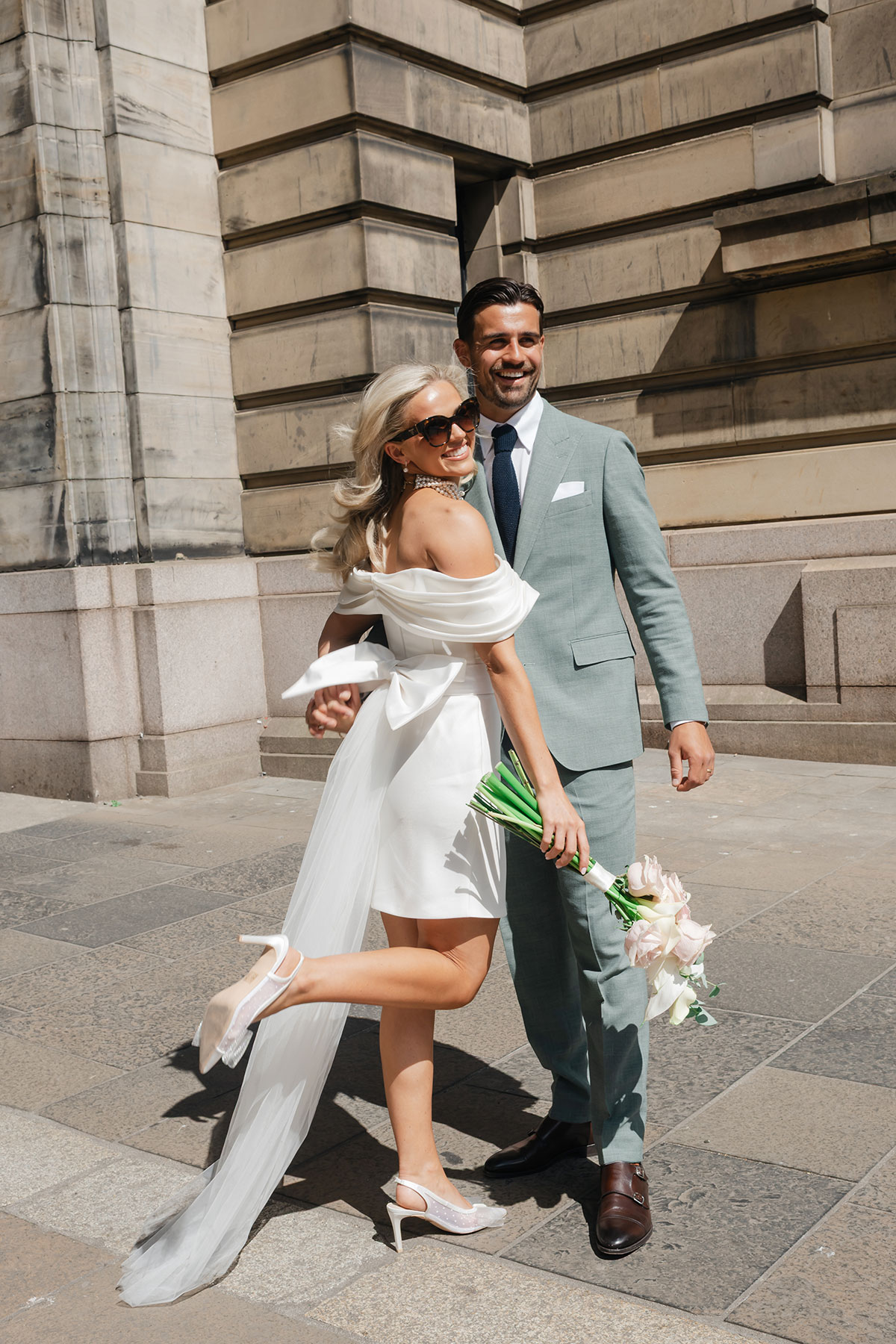 Bride wearing white mini wedding dress and pearl necklace smiles in sunglasses as groom in green suit stands beside her