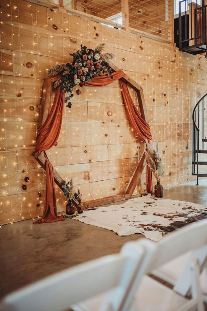 a hexagonal wooden arch with orange fabric draped over it and orange flowers on it leaning against a wooden wall with fairylights on it