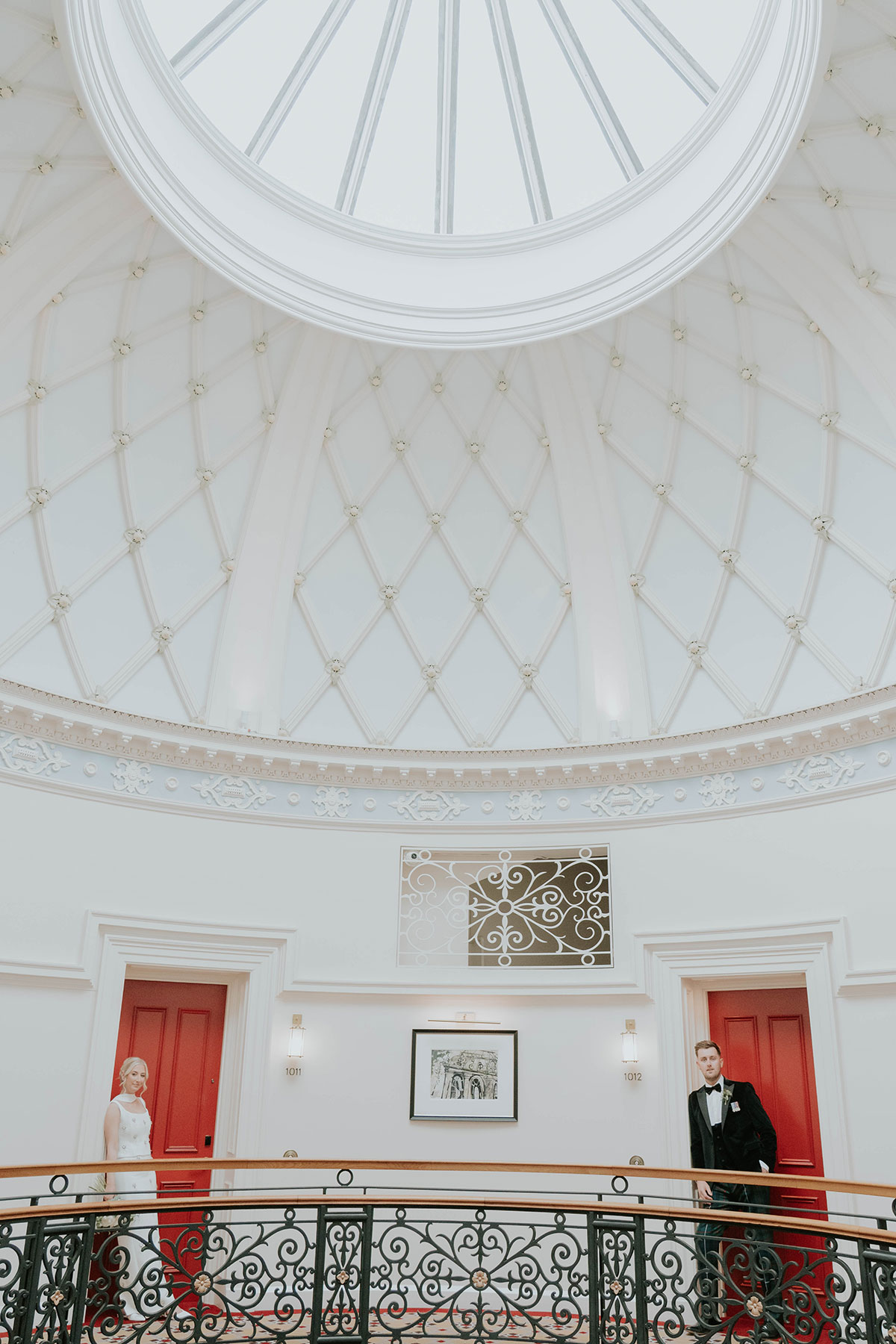 Bride and groom posing beneath a domed skylight and ornate iron balcony inside their Edinburgh wedding hotel