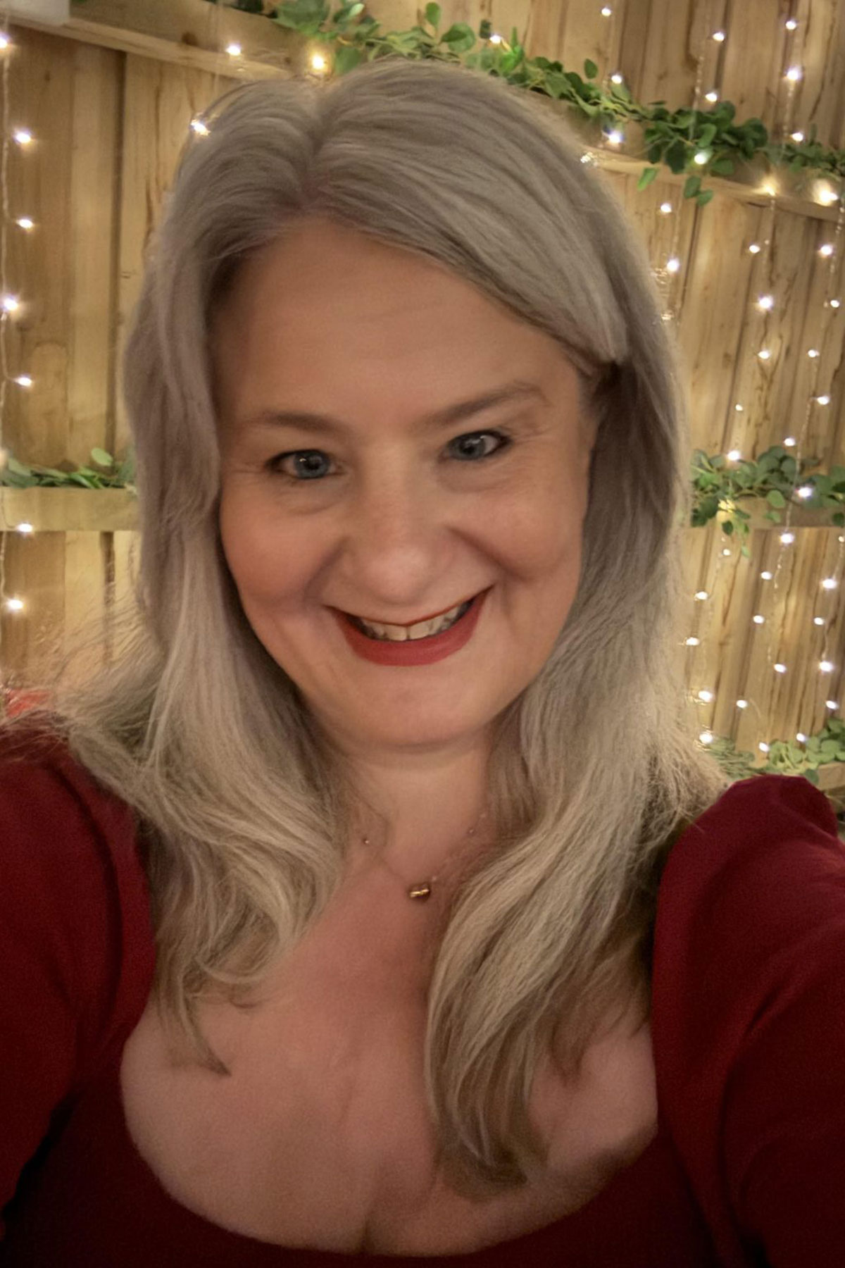 Female wedding supplier smiling in front of fairy light backdrop and greenery wall, warm indoor portrait