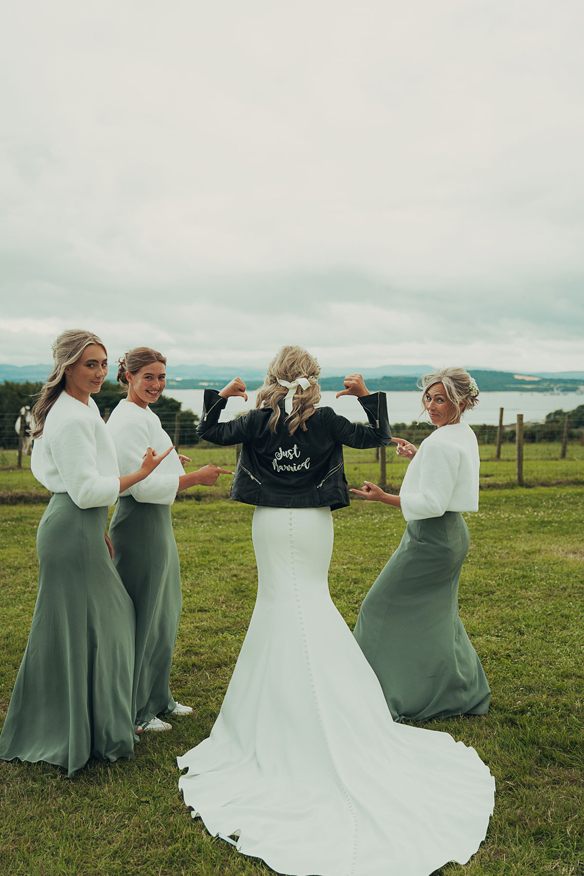 Bride wearing a black leather jacket with “just married” on the back stands with three bridesmaids in sage green dresses, all pointing towards her, with a coastal landscape in the background