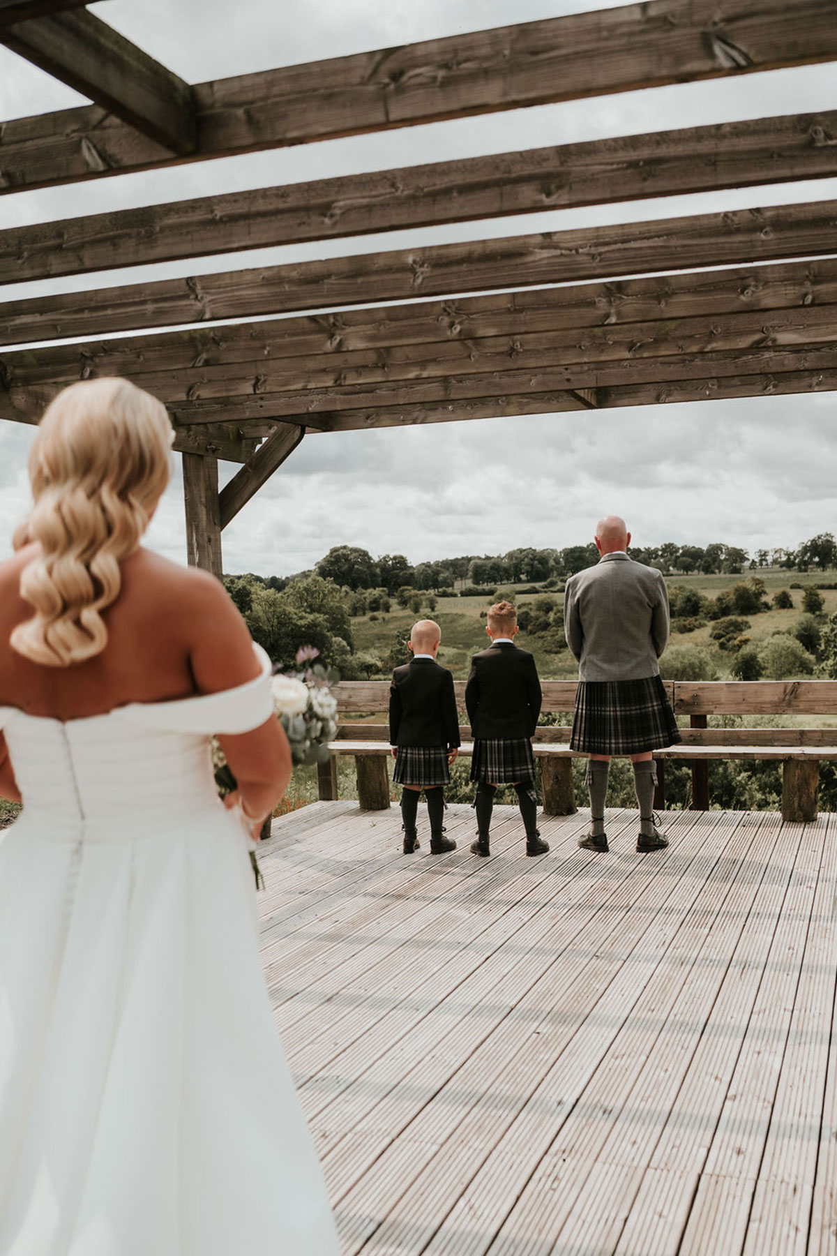 Bride walks towards the groom and two boys standing on a wooden deck overlooking the countryside.