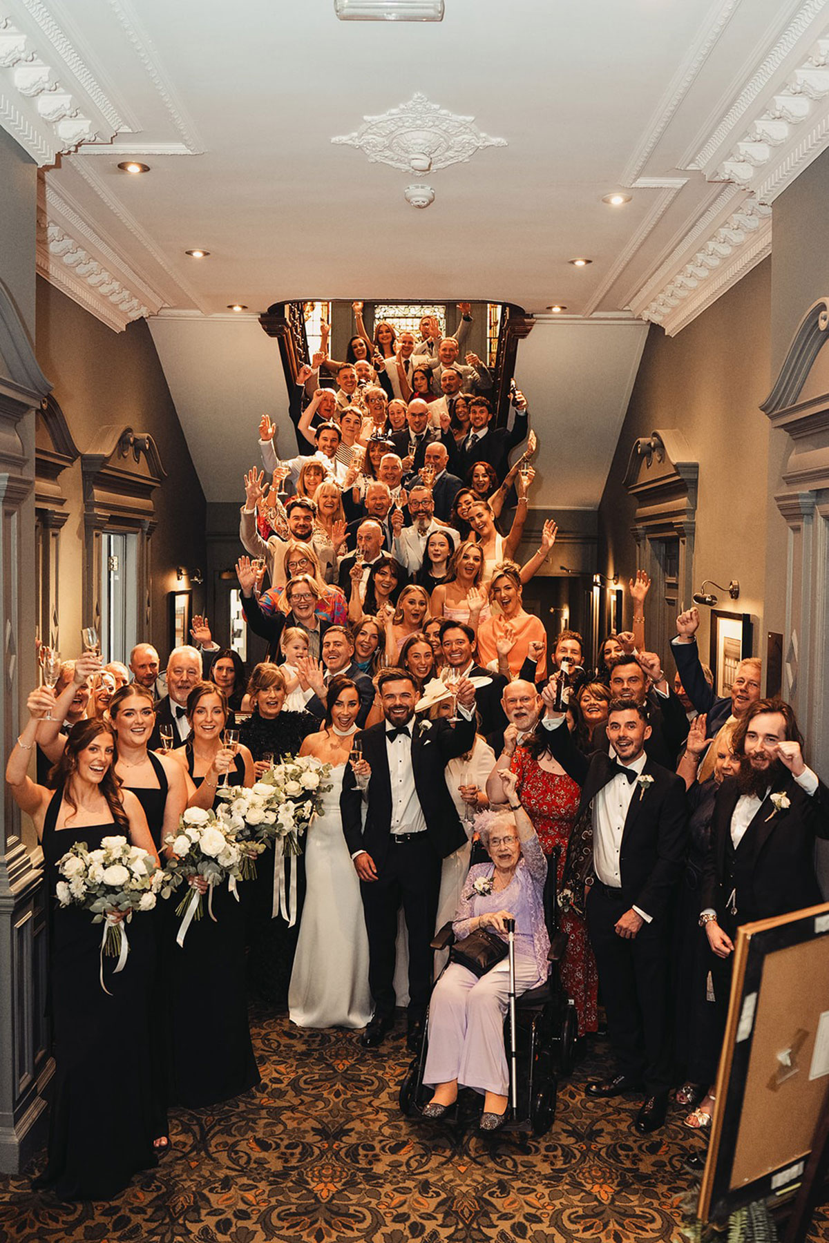 A full wedding party gathered on a grand staircase inside the hotel, raising glasses and cheering.