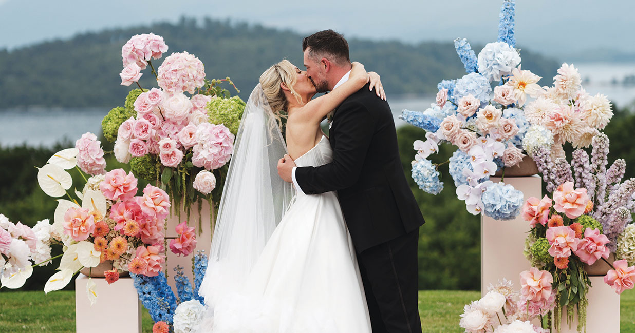 Bride and groom sharing a kiss outdoors beside colourful pink, blue and white floral installations, with loch views and rolling Scottish countryside in the background