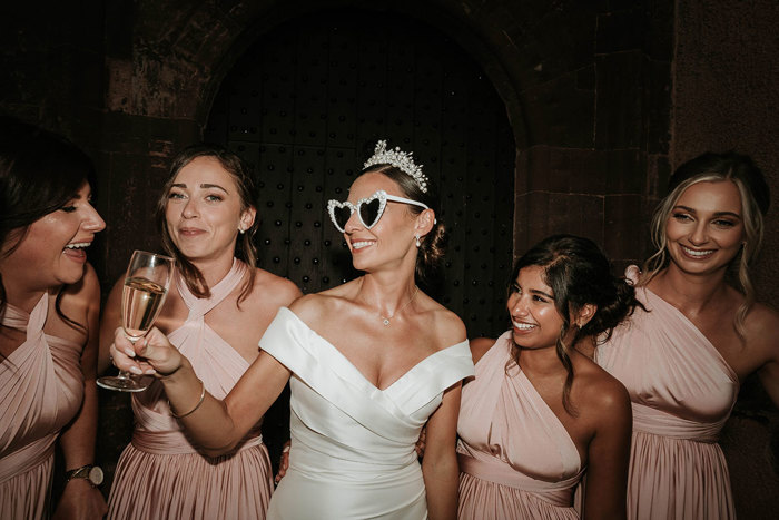 A bride in white heart sunglasses holds up a glass of champagne with two bridesmaids in pink dresses on either side of her