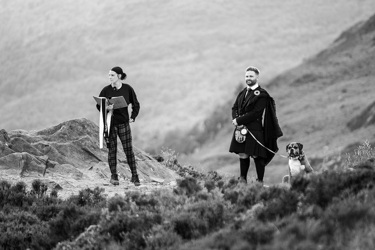 Groom in a traditional Scottish kilt stands beside his dog as a celebrant prepares for an outdoor mountain wedding ceremony in the Scottish Highlands