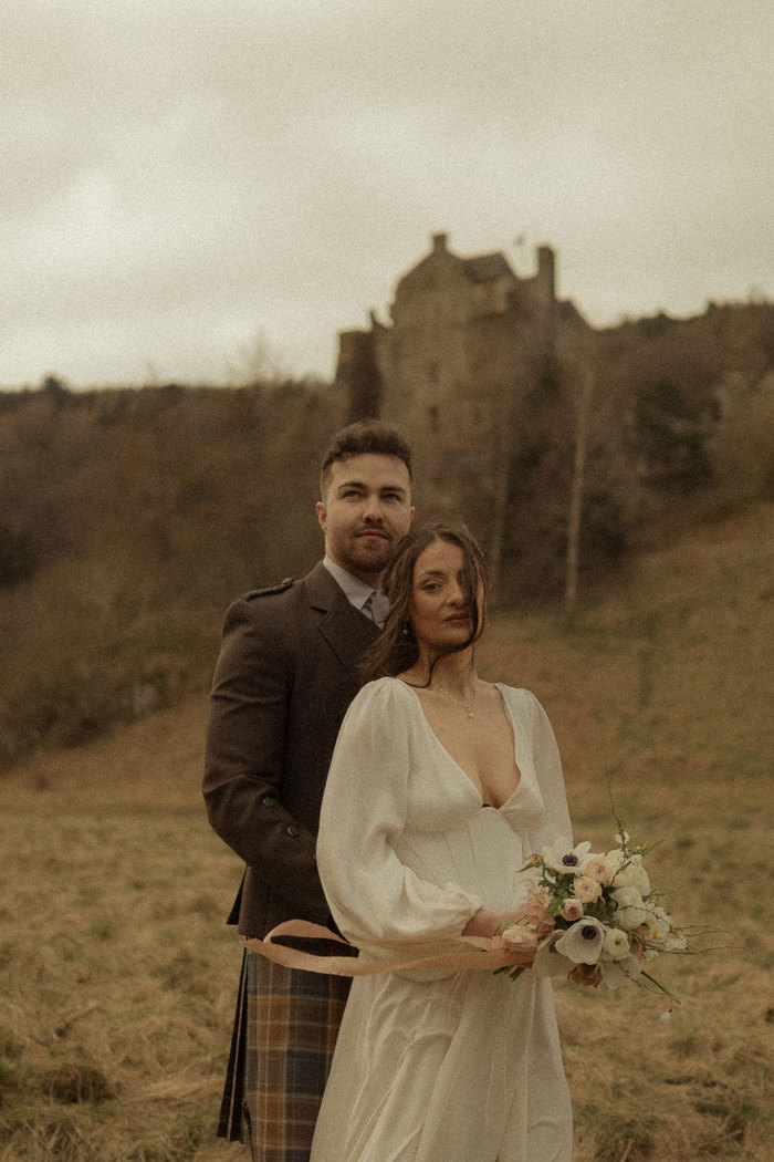 bride in plunging satin dress holding a bouquet of flowers, standing in front of groom in kilt outfit, stood outdoors on the grass with a small blurred castle in the background
