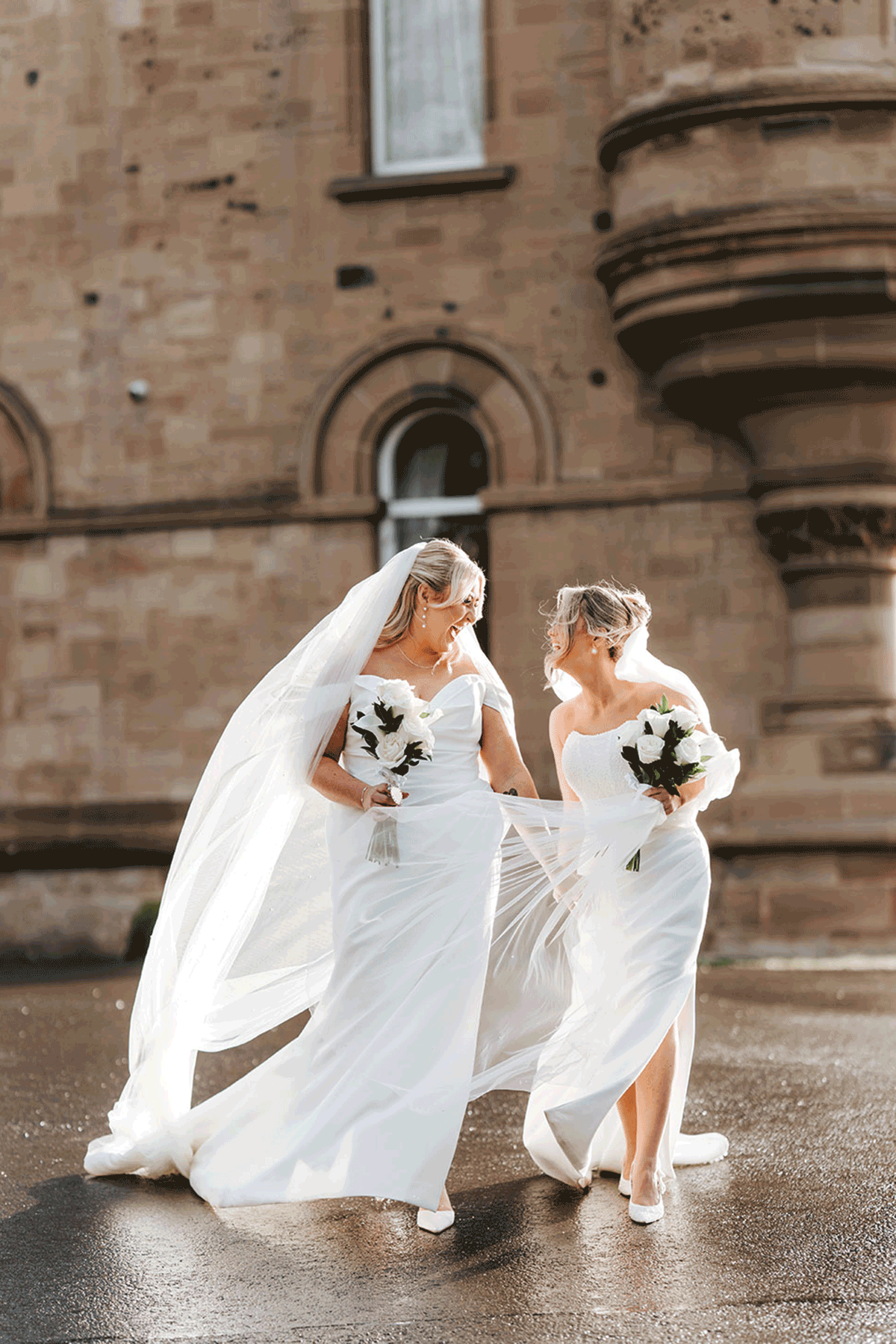 Two brides in elegant satin wedding dresses and long veils laughing together outside a historic Scottish venue after their ceremony