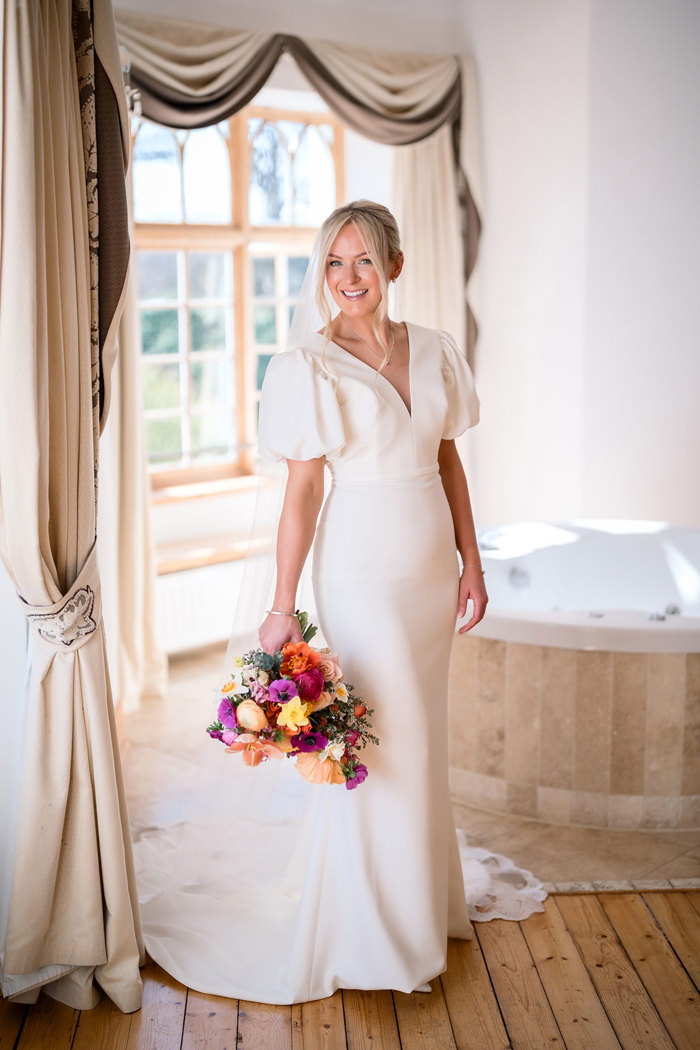 bride wearing a fitted wedding gown and puff sleeves holds a colourful bouquet at achnagairn castle