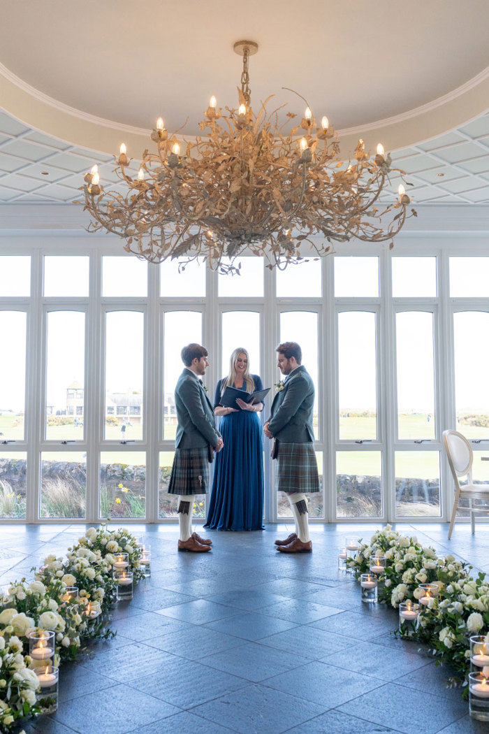 two grooms wearing green tweed kilt outfits standing in front of a person in a long blue dress. The windows in the background look out onto a golf course and there are rows of white flower decoration on a grey tiled floor