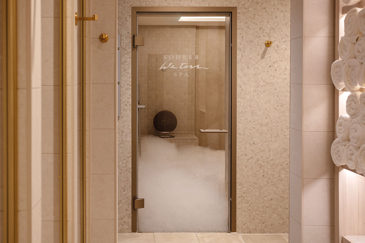 a nicely tiled room with gold fixtures and a glass door with the words 'Kohler Waters Spa' printed on and a hip height level of bubbles filling the room behind it