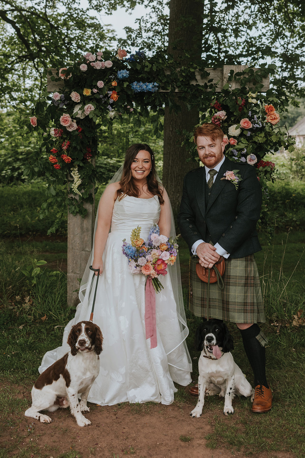 Bride and groom standing beneath a flower-covered wooden arch during an outdoor ceremony, holding a pastel bouquet with two dogs at their feet