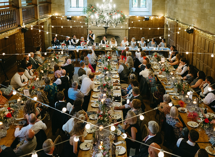 A room full of long wooden tables set for a meal with people sitting around them and fairylights above them 