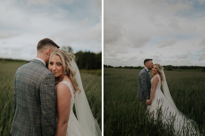 A Groom Wearing A Grey Checked Suit Nuzzles Into A Bride While Standing In A Field Of Tall Grass