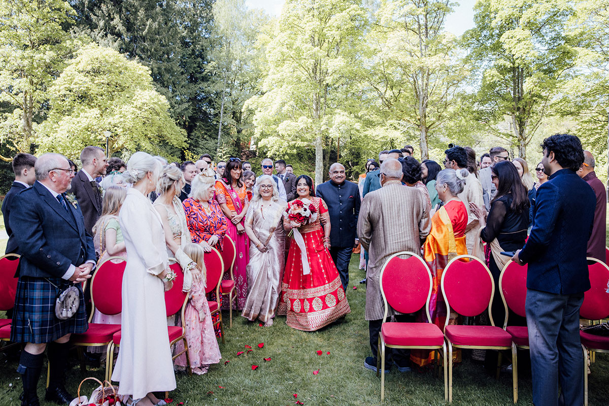 Bride walking down the aisle in a red Indian bridal outfit during an outdoor wedding ceremony at Atholl Palace Hotel