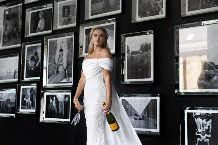 A bride wearing an off-the-shoulder fitted dress holding two glasses and a bottle of champagne