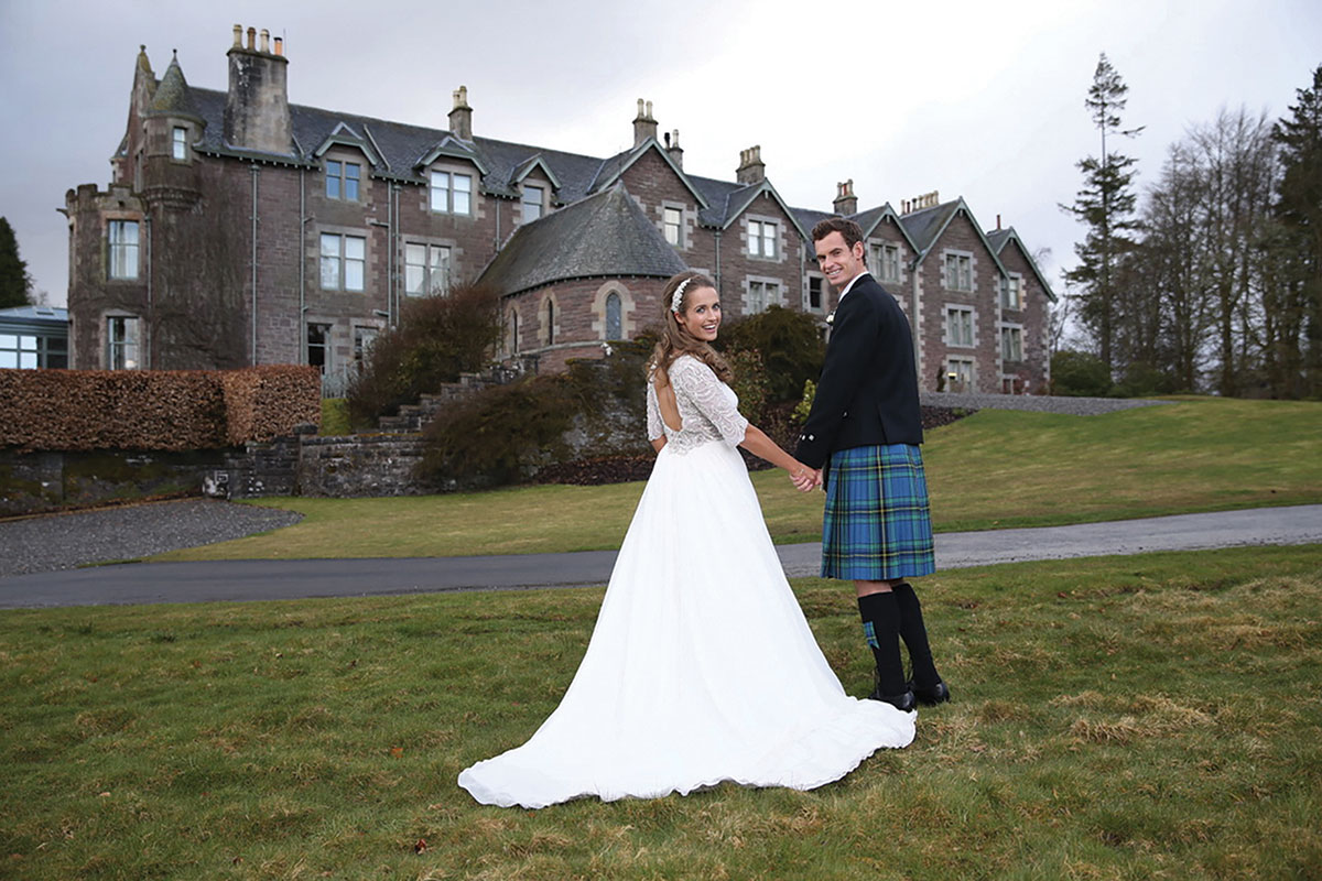 Bride and groom holding hands on the lawn in front of a grand Scottish castle venue; the bride wears a long-sleeved lace wedding dress and the groom wears a traditional kilt outfit
