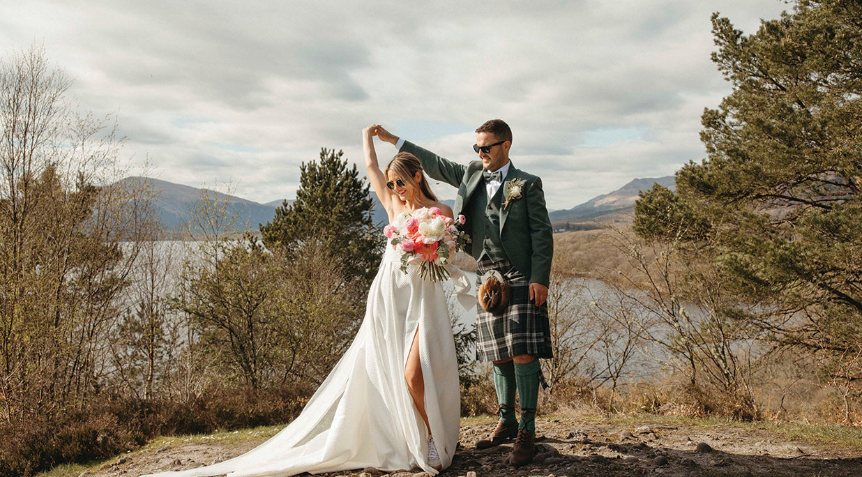A bride dances under a groom's arm. The bride is holding a large pink and white bouquet of flowers and wearing a textured wedding dress with skirt split. The groom is wearing a green tweed kilt outfit with bow tie. They are standing on stony ground with trees, hills and Loch Lomond in the background