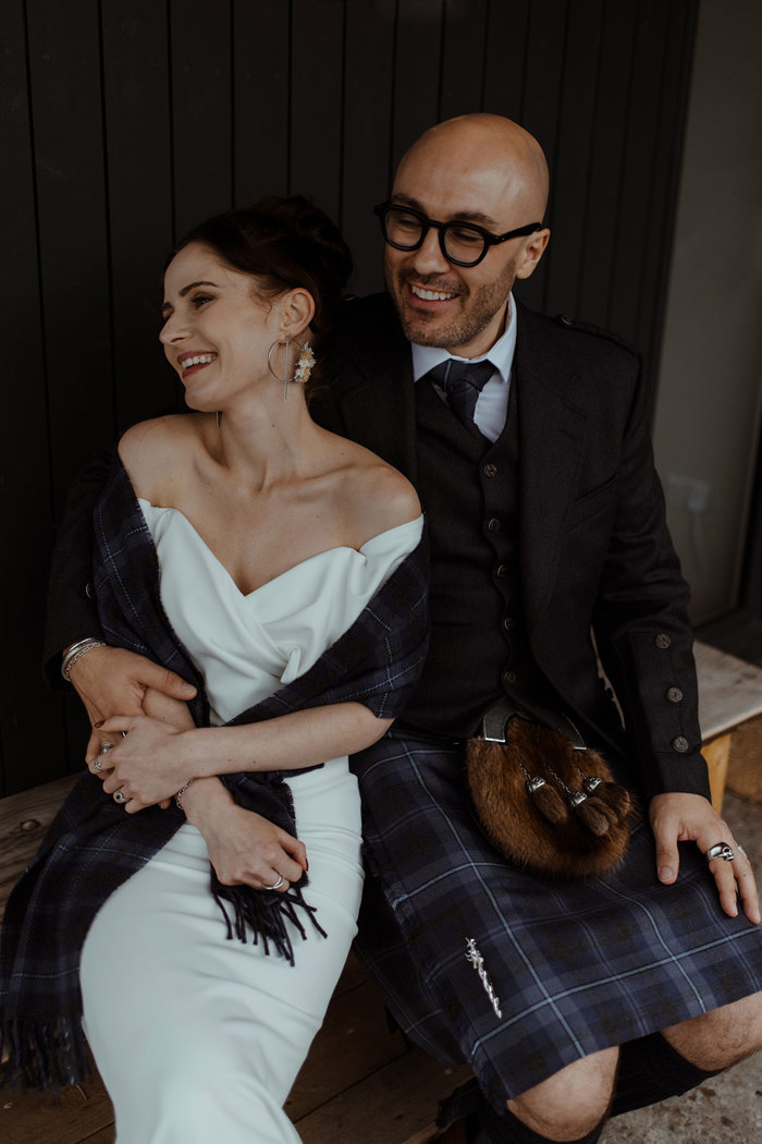 groom in blue, grey and white tartan kilt outfit and bride in off the shoulder white wedding dress and matching tartan shawl over her shoulders sit on wooden bench smiling side by side