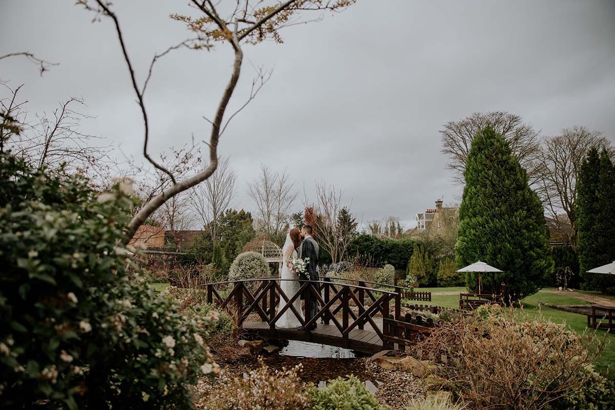bride and groom are stood kissing on small wooden bridge going over a stream between two sides of a green garden