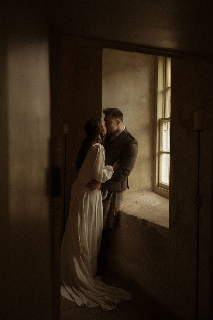 bride in trailing satin dress leans in, kissing groom in kilt outfit, leaning against stone windowsill in a shadowy room