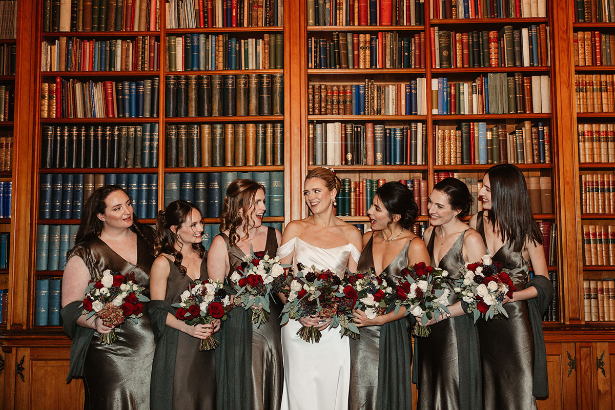 Bride and bridesmaids holding red-and-white bouquets, standing together in front of tall wooden bookshelves.