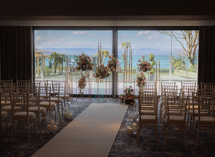 A room with wooden chairs set for a wedding ceremony with a white carpet down the middle facing a display of flowers and candles in front of windows and a view of the sea 
