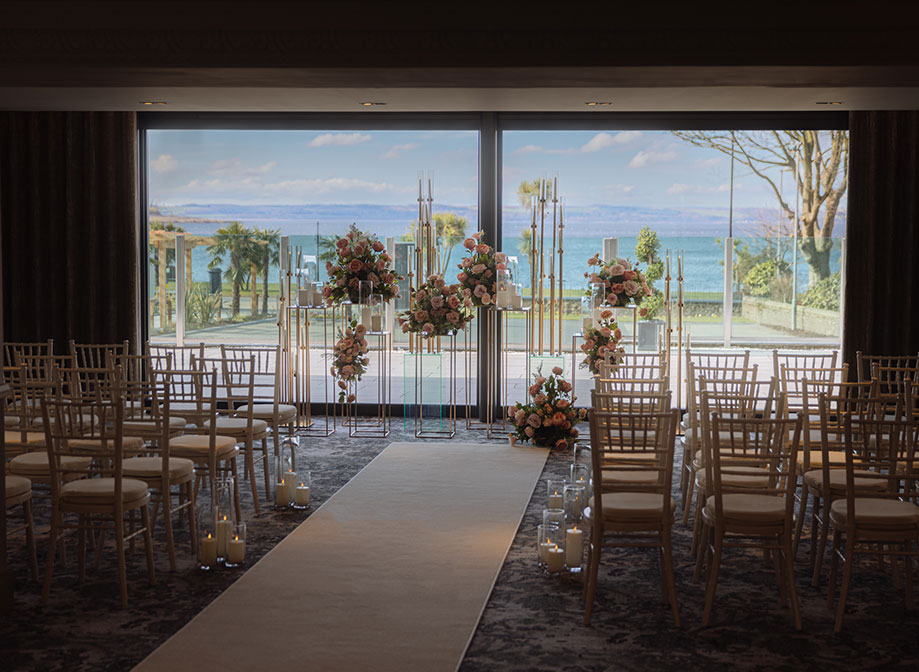 A room with wooden chairs set for a wedding ceremony with a white carpet down the middle facing a display of flowers and candles in front of windows and a view of the sea