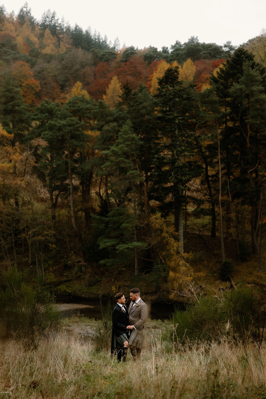 two grooms wearing kilts standing on grassy ground posing amid autumn trees
