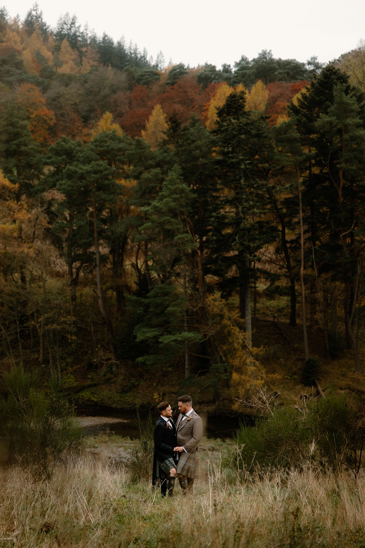 two grooms wearing kilts standing on grassy ground posing amid autumn trees