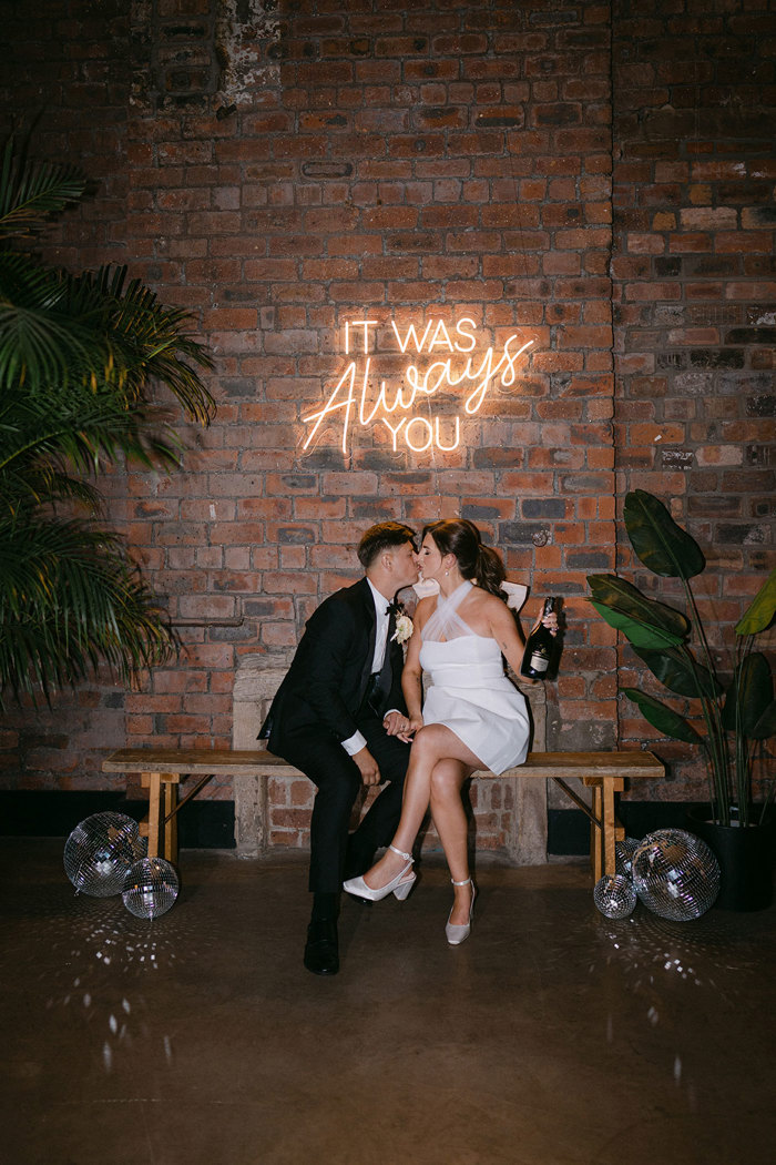 A bride and groom sitting on a bench kissing below a neon sign. There is a brick wall behind them and plants in pots either side on them.