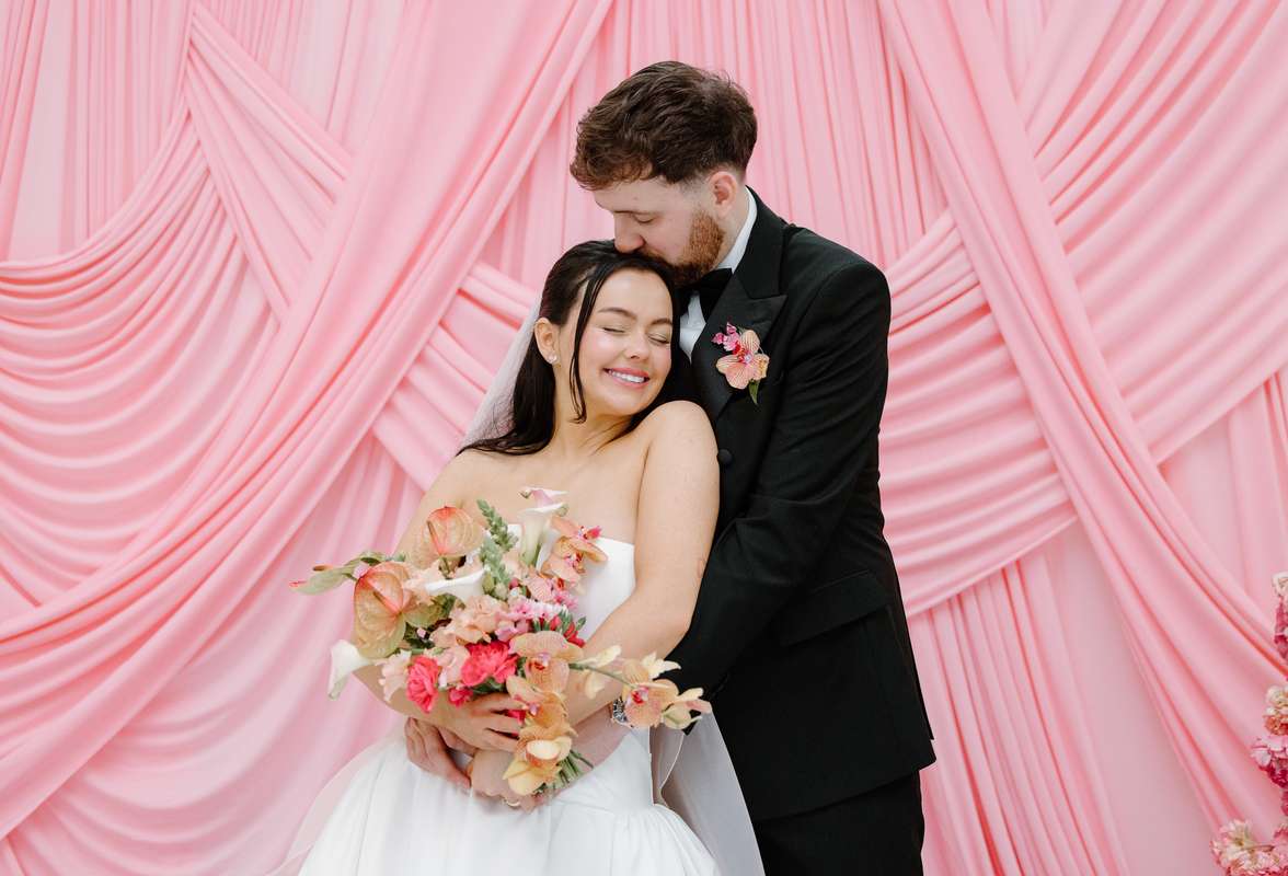 Bride and groom embrace with a colourful wedding bouquet in front of the pink draped ceremony backdrop at The Collector’s Hall Glasgow.