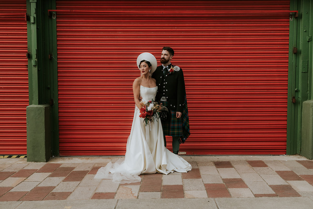 Bride and groom portrait against red shutter at BAaD Glasgow with groom in tartan kilt and bride holding bouquet