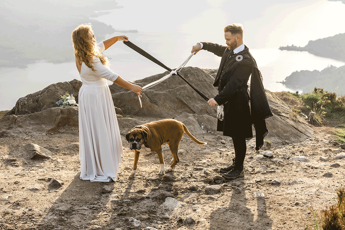 Couple perform a handfasting ceremony on a Scottish mountain summit while their boxer dog stands between them, with loch and hillside views in the background