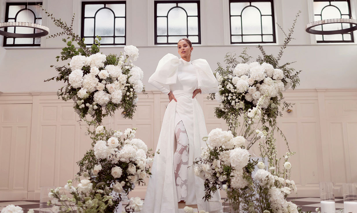 Emily Shak wearing an Opus Couture puff sleeve wedding dress at Glasgow wedding venue The Collector's Hall. She is surrounded by tall white flower arrangements.