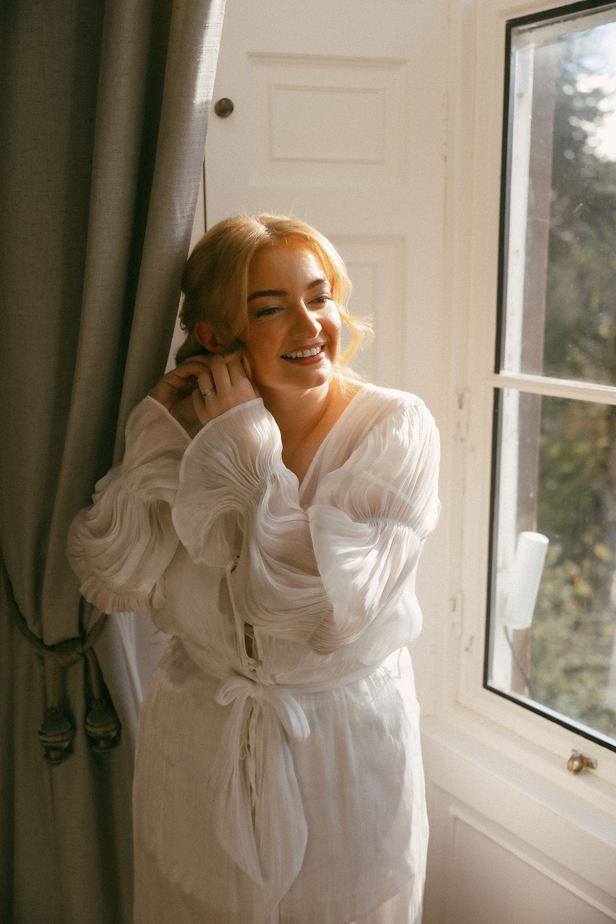 Bride getting ready by window in soft morning light wearing sheer robe on wedding morning in Scotland