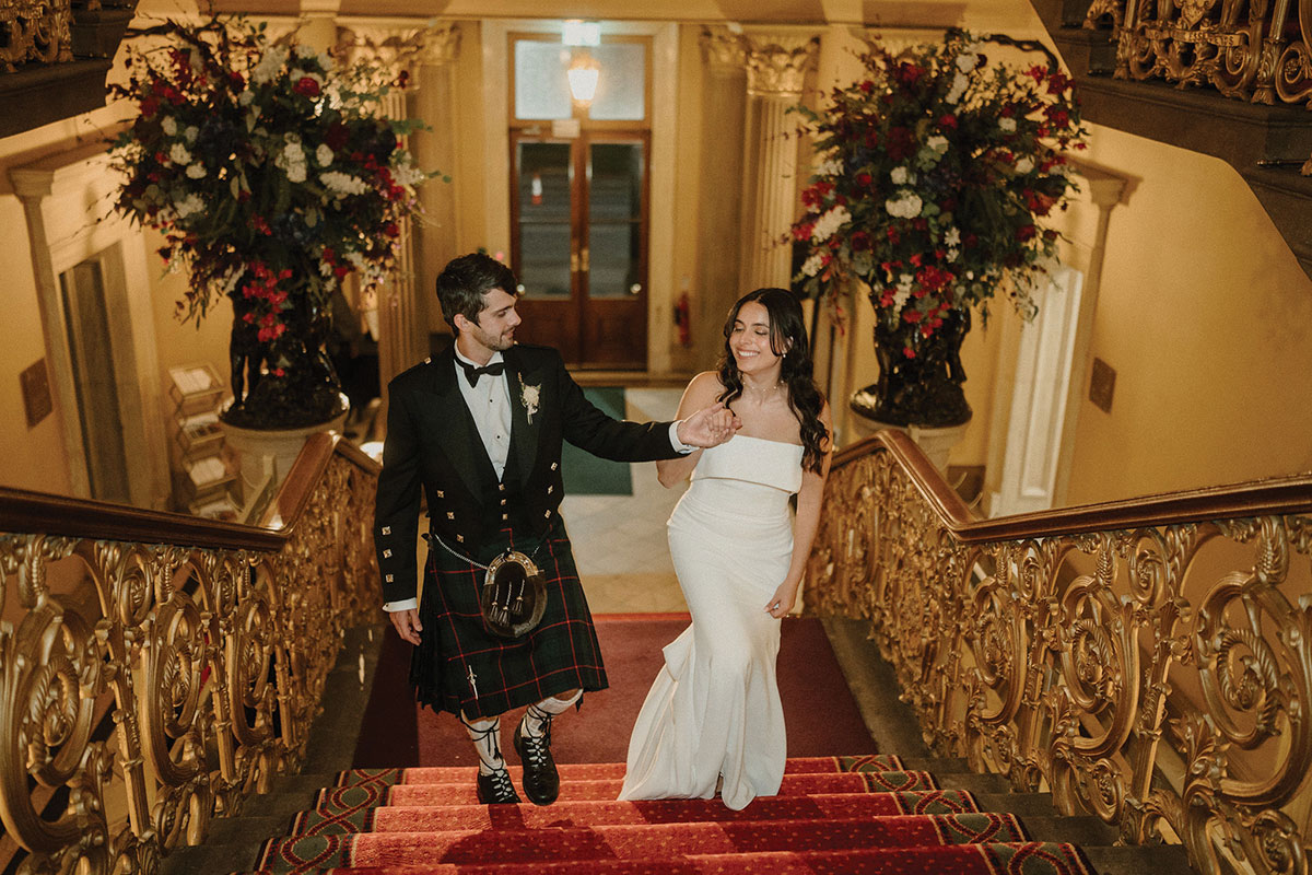 A bride and groom holding hands as they walk up the grand staircase at the Royal College of Physicians of Edinburgh, framed by ornate gold balustrades and floral arrangements