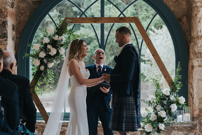 a bride and groom standing in front of a celebrant and an arched window during a wedding ceremony.