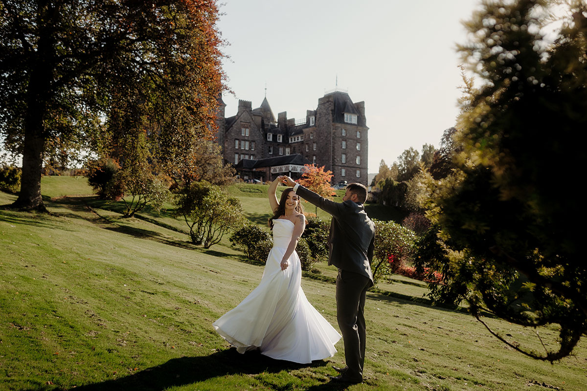 bride in wedding dress is spun around in dance by groom in suit on sunny grassy space outside brown building 