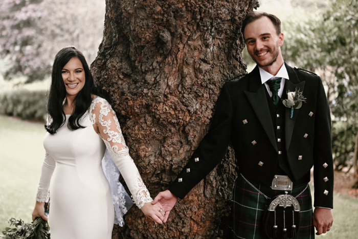 Bride and groom smile while they hold hands in front of a tree